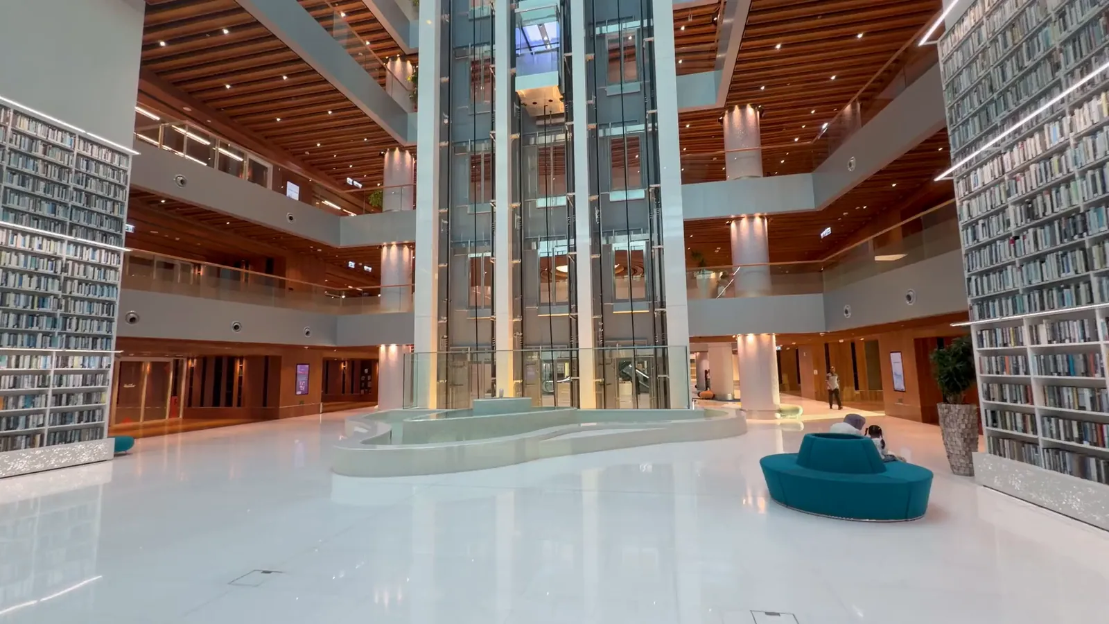 Modern library atrium with a glass elevator shaft rising between tiers of bookshelves